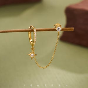 May include: Gold-toned earring set with a hoop, a star-shaped charm, and a chain connecting to a stud. The hoop is encrusted with small, clear stones. The star charm and stud feature a single, larger clear stone. The word "JEWSEEN" is at the bottom.