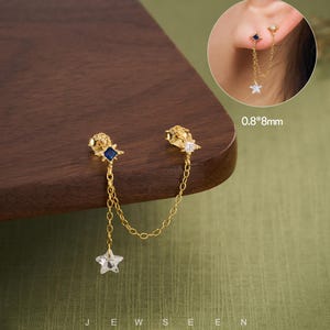 May include: Gold-coloured double earrings with star and square-shaped accents. One earring has a blue square, the other a clear star. A delicate chain connects the two earrings. The earrings are displayed on a wooden surface.