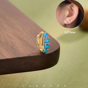 May include: A gold-colored hoop earring with five blue opal stones. The earring is shown on a wooden surface. The text "JEW SEEN" is at the bottom of the image. A smaller image shows the earring in use. The earring measures 0.8*6mm.