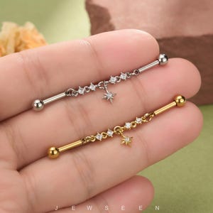 May include: Two industrial barbell piercings, one silver-toned and one gold-toned, each featuring a chain with small clear stones and a central star-shaped charm. The barbells have spherical ends and are held in a hand against a green background.