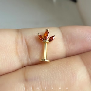 May include: A gold-tone flat back stud earring featuring a vertical post and a decorative top element shaped like a three-petaled flower, adorned with three marquise-cut orange gemstones arranged in a fan-like pattern at the top of the post.