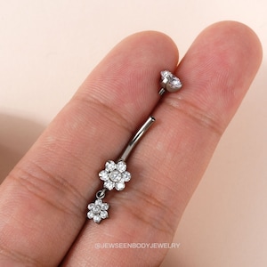 Puede incluir: Un anillo de ombligo plateado con dos colgantes de flores, cada uno adornado con piedras de circonita cúbica transparentes.