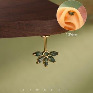 Op de afbeelding: Een goudkleurige, bloemvormige oorbel met groene steenaccenten. De oorbel hangt aan een gouden staaf. De tekst "JEWSEEN" staat onderaan de afbeelding. De oorbel meet 1,2 mm x 6 mm.