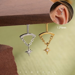 Puede incluir: Dos pendientes tipo barbell con una medida de 1,2*6mm, uno plateado y otro dorado. Cada uno presenta una barra curva conectada por una cadena a un dije en forma de estrella con una piedra clara central. Un recuadro circular muestra el pendiente puesto.