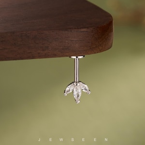 May include: A silver earring with a floral design, adorned with clear gemstones. The earring hangs from a dark brown wooden surface. The word "JEWSEEN" is visible at the bottom of the image.