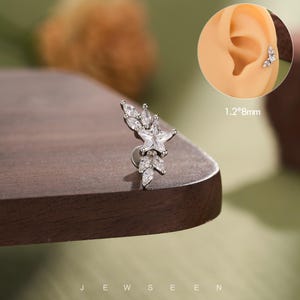 May include: A silver-toned stud earring featuring a central star-shaped clear gemstone surrounded by marquise-cut clear stones in a leaf-like arrangement. The piece is labeled with the dimensions 1.2*8mm and is shown inserted into an ear.