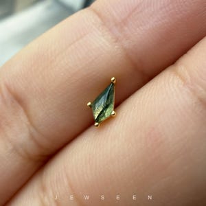 May include: A kite-shaped moss agate gemstone set in a gold prong setting. The stone features translucent green hues with dark, branch-like inclusions throughout. The piece is small and delicate, showcasing a geometric cut with four gold prongs.