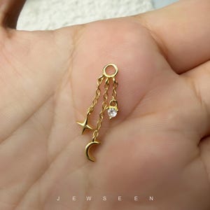 May include: A gold-toned charm featuring a small circular loop at the top, from which three delicate chains descend. One chain ends in a four-pointed star, another in a crescent moon, and the third in a single clear, round-cut gemstone.