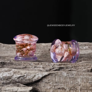 May include: Two purple acrylic ear gauges with pink flowers encased in clear resin. The gauges are on a wooden surface. @JEWSEENBODYJEWELRY
