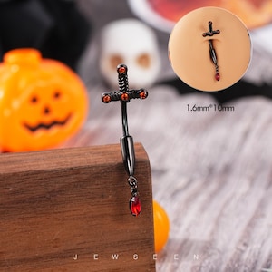 May include: A black sword-shaped belly button ring with red accents and a dangling red gem. The ring is 1.6mm x 10mm. The word "JEWSEE" is printed at the bottom. The image also includes a carved orange pumpkin and a white skull.