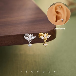 May include: Two stud earrings with a floral design, one silver-toned and one gold-toned, each featuring clear, sparkling gemstones. The earrings are shown with a close-up of an ear piercing. The text "JEWSEEN" is at the bottom.