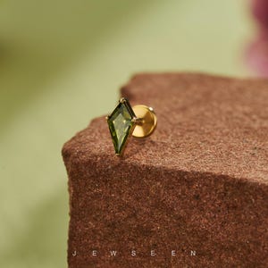 May include: A gold stud earring featuring a green, diamond-shaped gemstone. The earring is set on a brown surface with a green background. The word "JEWSEN" is printed at the bottom of the image.