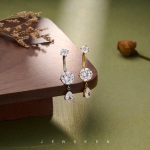 Puede incluir: Dos piercings para el ombligo con piedras preciosas claras y brillantes. Uno es plateado, el otro dorado. Cada uno tiene una gema redonda grande, una gema superior más pequeña y una gema en forma de lágrima. La palabra "JEWSEEN" está en la parte inferior.