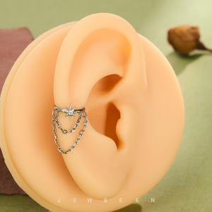 May include: A silver ear cuff with a starburst design and a clear central stone, plus dangling chains. The cuff is displayed on a light-toned ear model. The word "JEWSEEN" is visible at the bottom.