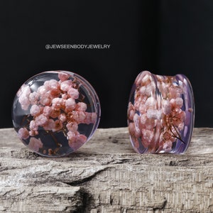 May include: Two clear acrylic ear gauges with pink pressed flowers inside. The gauges are on a wooden surface.