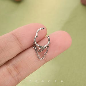 May include: Silver septum ring with a dangling chain design. The jewellery features a hinged closure and a small decorative element at the top of the chain. The word "JEWSEEN" is visible at the bottom of the image.