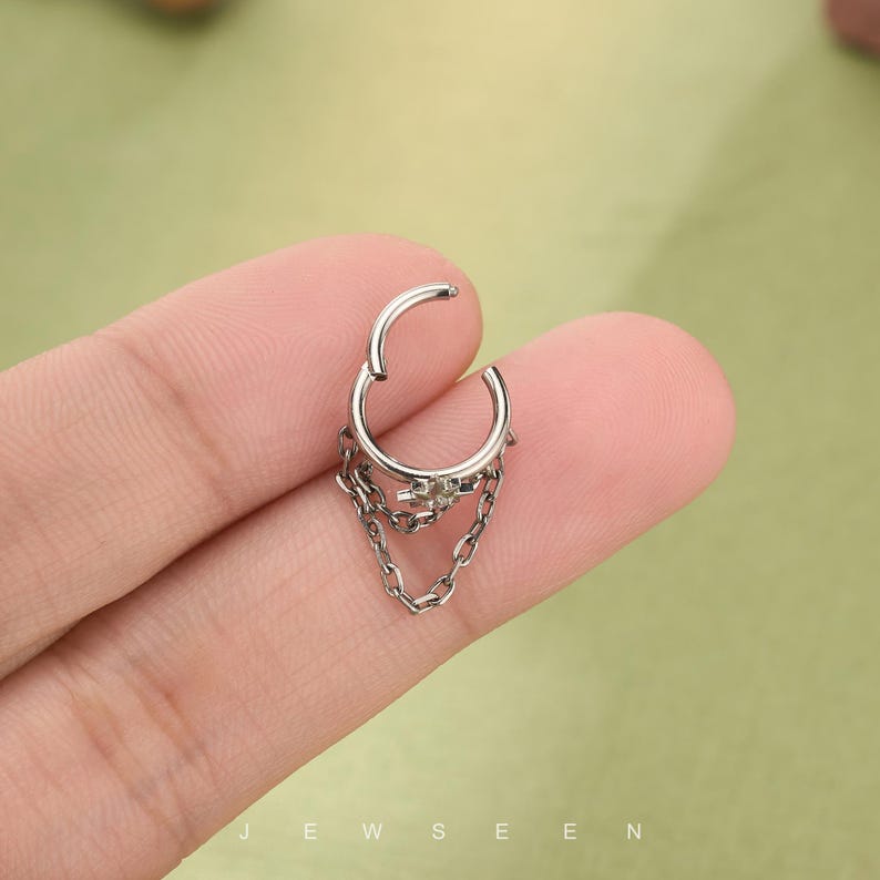 May include: Silver septum ring with a dangling chain design. The jewellery features a hinged closure and a small decorative element at the top of the chain. The word "JEWSEEN" is visible at the bottom of the image.