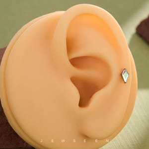 May include: A kite-shaped stud earring featuring a multifaceted iridescent crystal set in a dark metal frame, positioned on the cartilage of a beige ear model. The text JEWSEEN is printed in white at the bottom of the display base.