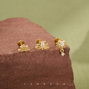 Puede incluir: Tres pendientes de botón dorados con diseños de flores de loto, cada uno con una piedra preciosa transparente central. Un pendiente tiene una piedra preciosa colgante. Los pendientes se exhiben sobre una superficie marrón con la palabra "JEWSEEN" en la parte inferior.