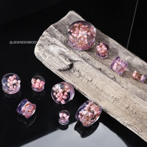 May include: A set of eight pink and clear glass ear gauges with embedded pink flowers. The gauges are arranged on a piece of weathered wood.