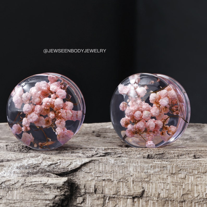 May include: Two clear glass ear gauges with pink pressed flowers inside. The gauges are on a wooden surface. @Jewseenbodyjewelry
