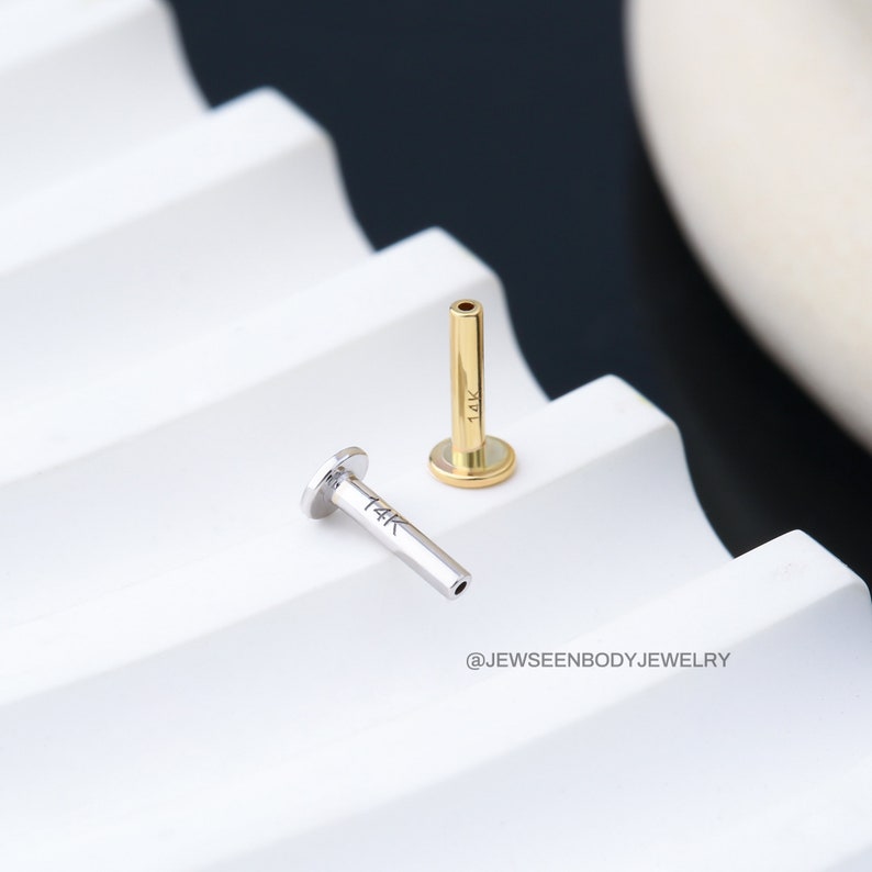 Op de afbeelding: Twee 14 karaats gouden lichaamsieraden, een in goud en een in zilver, worden getoond op een wit, getextureerd oppervlak. De sieraden zijn beide recht en hebben een platte bovenkant en een holle onderkant.