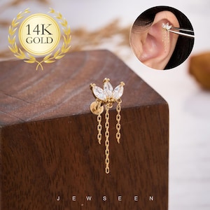 May include: A gold-colored earring with a floral design, featuring three clear, sparkling stones and dangling chains. The earring is displayed on a wooden block. The text "14K GOLD" is visible in a circular emblem.