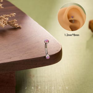 May include: A curved eyebrow piercing with pink opal-like stones at each end. The jewelry is made of silver-colored metal. The image includes a close-up of the piercing in an ear, and the text "1.2mm*8mm".