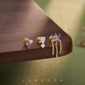 Może przedstawiać: Trzy złote kolczyki z akcentami z kamieni szlachetnych są prezentowane na drewnianej powierzchni. Jeden kolczyk ma fioletowe i białe kamienie, drugi ma biały kamień w kształcie gruszki, a trzeci fioletowy kwiatowy wzór z wiszącymi łańcuszkami. Słowo "JEWSEEN" znajduje się na dole.