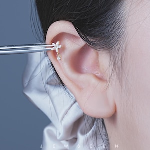 Op de afbeelding: Een delicate goudkleurige oorbel met een klein bloemmotief met heldere, sprankelende kristallen. Een enkele, druppelvormige kristal bungelt eronder. De oorbel wordt door een pincet bij een oor gehouden.
