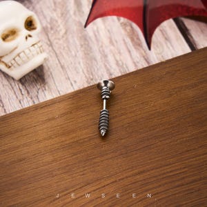 May include: A silver-colored stud earring shaped like a screw, with a detailed spiral design on the lower portion. The earring is set against a dark wooden surface. The word "JEWSEEN" is visible at the bottom.