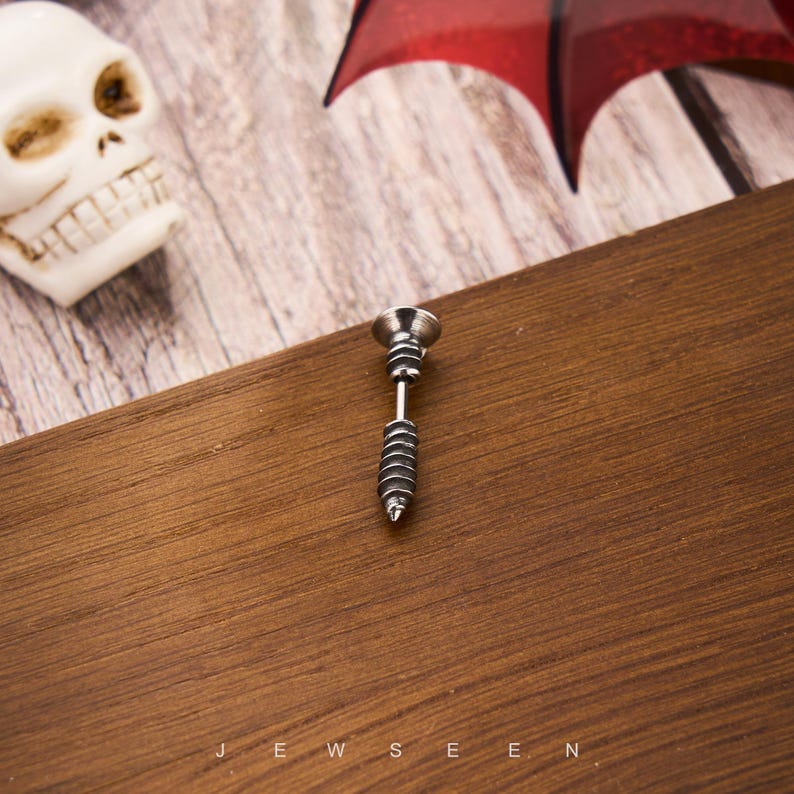 May include: A silver-colored stud earring shaped like a screw, with a detailed spiral design on the lower portion. The earring is set against a dark wooden surface. The word "JEWSEEN" is visible at the bottom.
