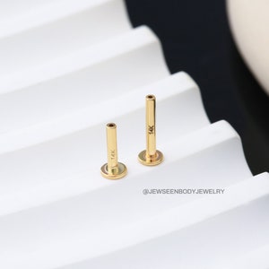 Op de afbeelding: Twee gouden 14 karaat barbell-studs, een langer dan de andere, op een wit oppervlak.  @Jewseenbodyjewelry