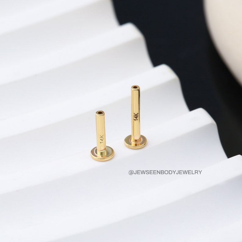 Op de afbeelding: Twee gouden 14 karaat barbell-studs, een langer dan de andere, op een wit oppervlak.  @Jewseenbodyjewelry