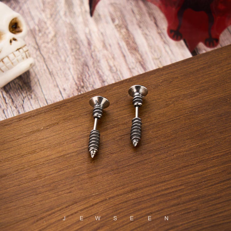 May include: A pair of silver-colored screw-shaped earrings. Each earring features a detailed design mimicking a screw, with a head, a threaded body, and a pointed tip. The earrings are displayed on a wooden surface.