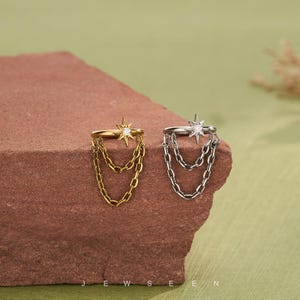 May include: Two ear cuffs, one gold and one silver, each with a starburst design and chain dangles. They are presented on a textured, reddish-brown surface against a soft green backdrop. The word "JEWSEN" is printed at the bottom.