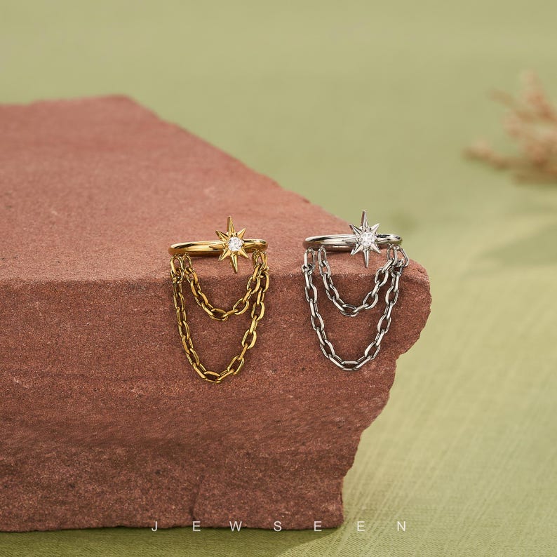 May include: Two ear cuffs, one gold and one silver, each with a starburst design and chain dangles. They are presented on a textured, reddish-brown surface against a soft green backdrop. The word "JEWSEN" is printed at the bottom.