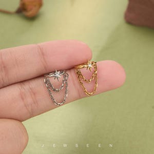 May include: Two ear cuffs, one silver and one gold, each with a star design, a central gem, and dangling chain details. The word "JEWSEEN" is printed at the bottom of the image. Jewellery.