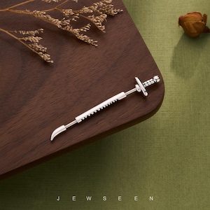 Puede incluir: Una joya plateada con forma de espada, con una hoja curva y un borde dentado. La joya tiene una empuñadura en forma de cruz y se exhibe sobre una superficie de madera oscura. La palabra "JEWSEEN" está impresa en la parte inferior.