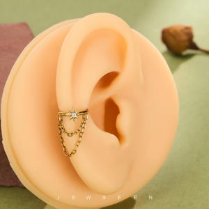 May include: Gold ear cuff with a starburst design and a central clear stone, plus dangling chain details. The cuff is displayed on a light-toned ear model. The word "JEWSEEN" is visible at the bottom of the image.