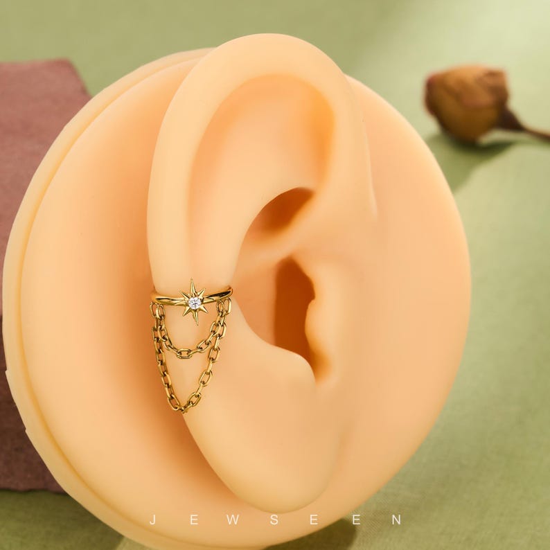 May include: Gold ear cuff with a starburst design and a central clear stone, plus dangling chain details. The cuff is displayed on a light-toned ear model. The word "JEWSEEN" is visible at the bottom of the image.