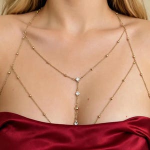 May include: A gold-toned body chain necklace featuring delicate link chains with small evenly spaced beads. The design includes a central vertical drop element adorned with three clear round-cut stones, creating a layered look that drapes across the chest area.