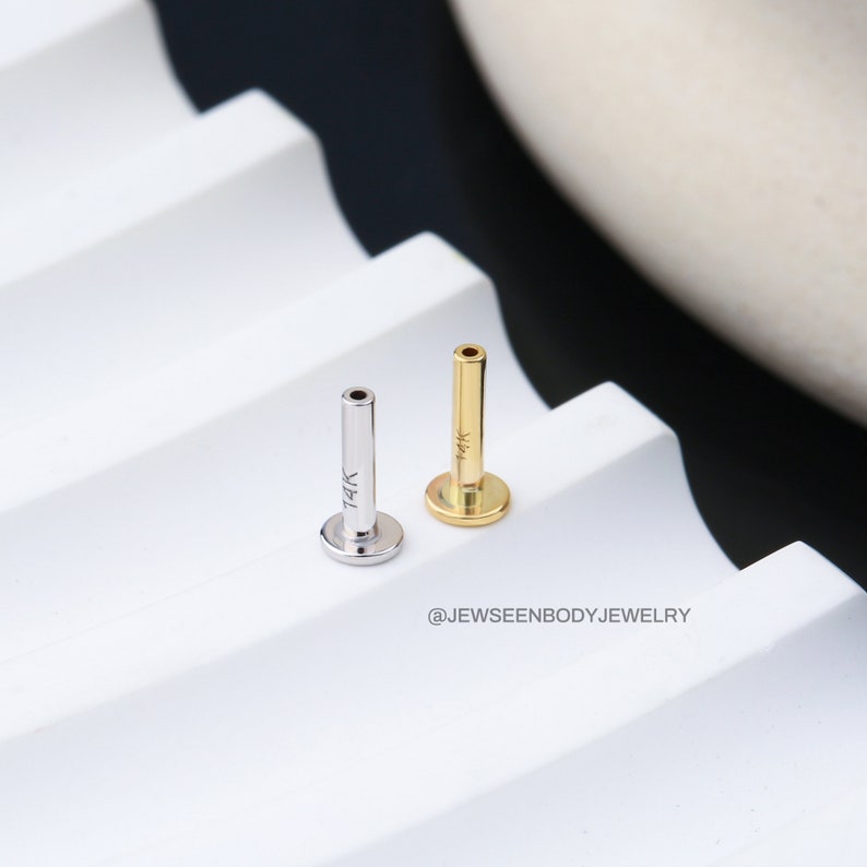Op de afbeelding: Twee 14 karaats gouden lichaamsieraden, een in zilver en een in goud, worden getoond op een wit oppervlak. De sieraden hebben de vorm van een kleine, rechte staaf met een platte schijf aan het einde. De zilveren staaf is gemarkeerd met "14K" en de gouden staaf is gemarkeerd met "14K".