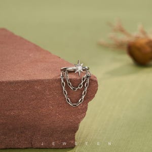 May include: A silver ear cuff with a starburst design and a central gemstone, plus dangling chain details. The jewellery is shown on a textured, reddish-brown surface against a green backdrop. The word "JEWSEEN" is printed at the bottom.