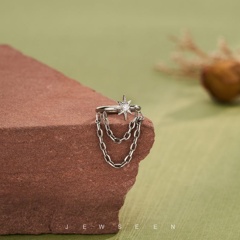 May include: A silver ear cuff with a starburst design and a central gemstone, plus dangling chain details. The jewellery is shown on a textured, reddish-brown surface against a green backdrop. The word "JEWSEEN" is printed at the bottom.