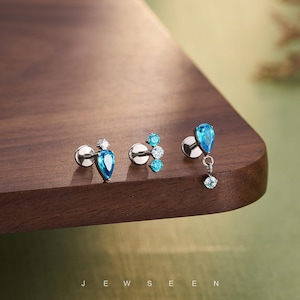 May include: Three pairs of silver earrings with blue and clear gemstones. The earrings are displayed on a dark wooden surface. The text "JEW SEEN" is visible at the bottom of the image.