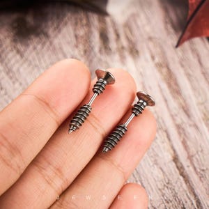 May include: A pair of silver-colored screw-shaped earrings. Each earring has a flat, circular head and a threaded shaft, resembling a miniature screw. The earrings are displayed against a blurred background.