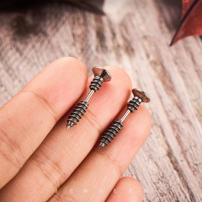 May include: A pair of silver-colored screw-shaped earrings. Each earring has a flat, circular head and a threaded shaft, resembling a miniature screw. The earrings are displayed against a blurred background.