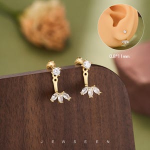 Könnte beinhalten: Ein Paar goldfarbene Ohrringe mit einem runden, klaren Stein als Stecker, der mit einem hängenden Element mit drei klaren, marquise-geschliffenen Steinen verbunden ist. Die Ohrringe messen 0,8*11mm. Ein Inset zeigt den Ohrring am Ohrläppchen.