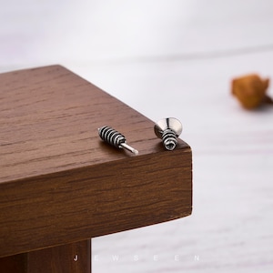 May include: Two silver-colored screw-shaped earrings. One earring has a cylindrical head with horizontal grooves, and the other has a flat, circular head. The earrings are displayed on a dark brown wooden surface.
